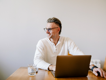 Ein Mann sitzt vor seinem Laptop und blickt lachend zur Seite.